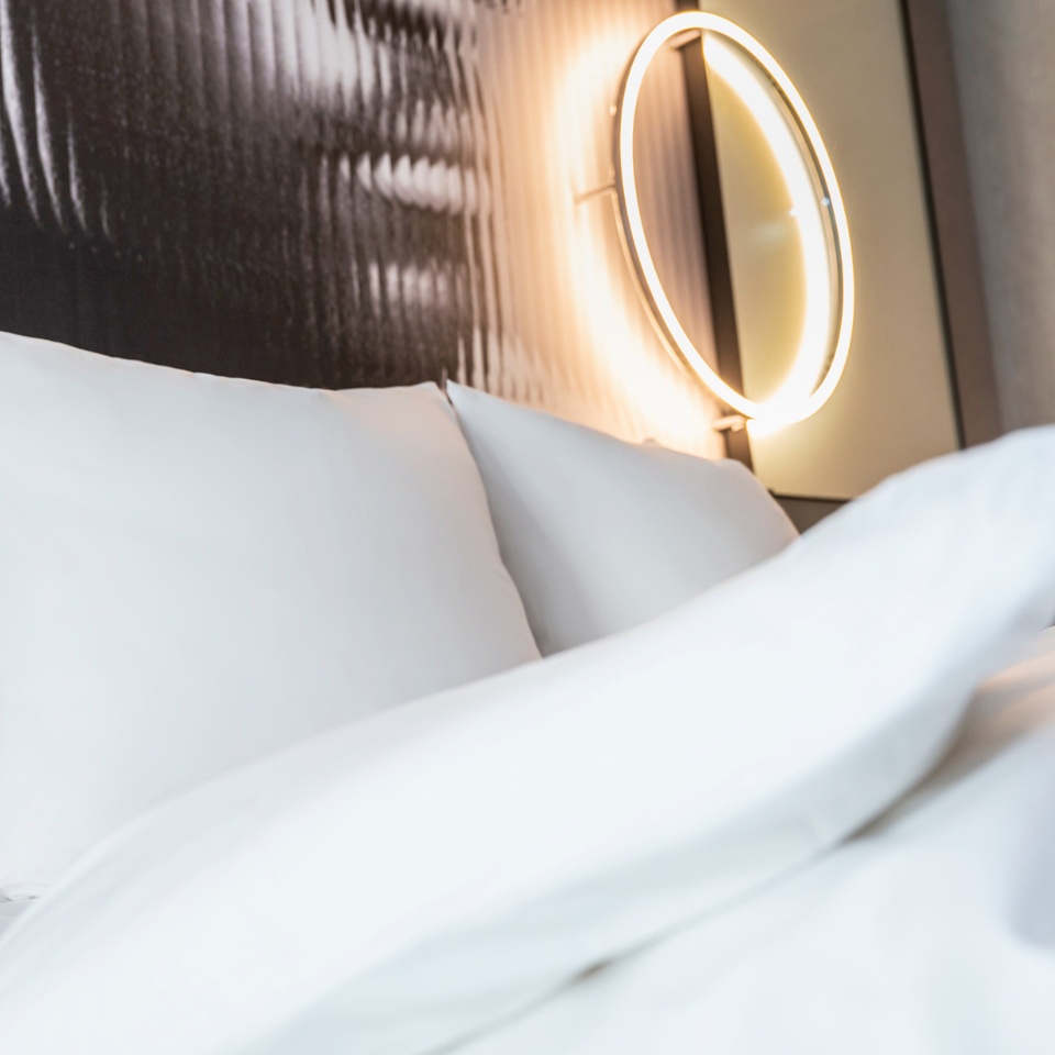 Close-up of a double bed with white linens, next to it is a lamp in form of a ring on the wall