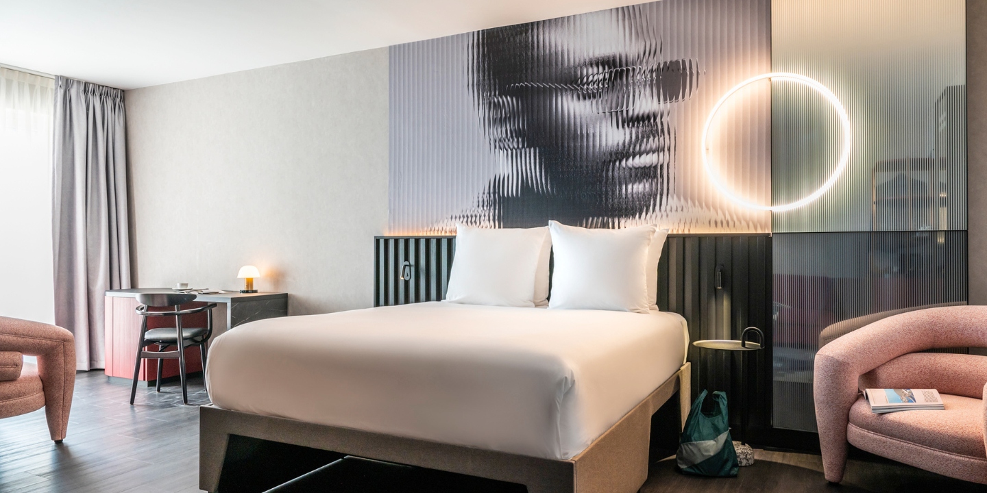Guest room with a double bed, a pink chair and a small desk in front of an artwork on the wall