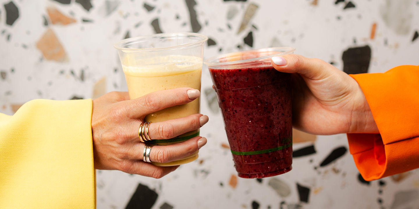 Two hands toast with smoothies in clear cups; one is yellow, the other red. The hands wear colorful sleeves, against a speckled background, conveying a cheerful mood.