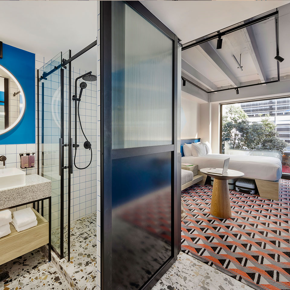 Modern hotel room with geometric decor: features a glass shower with black fixtures, a round mirror, a bed by a large window, and a patterned rug. Bright and inviting.