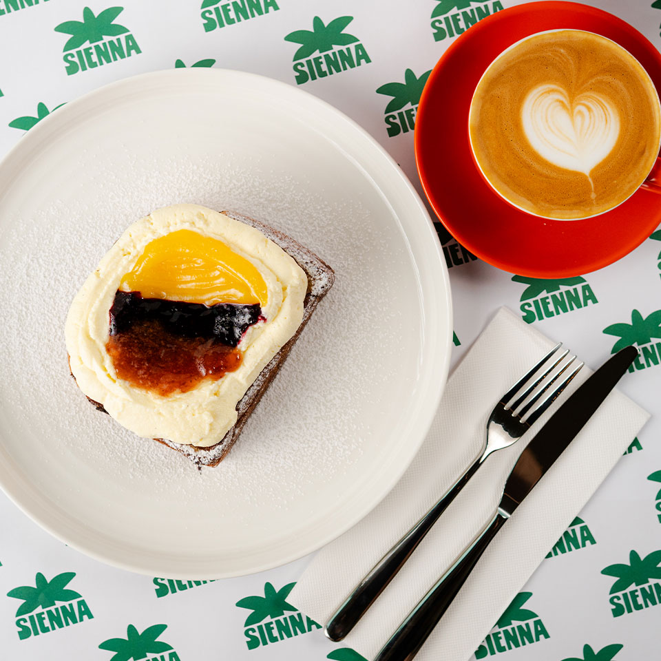 A dessert topped with cream, peach, and berry jam on a white plate, next to a cappuccino with heart art. Fork and knife on a napkin nearby.