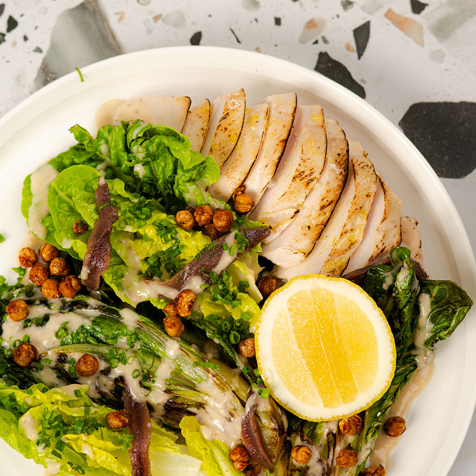 A colorful Caesar salad features sliced grilled chicken, romaine lettuce, crispy chickpeas, anchovies, and a lemon wedge, drizzled with creamy dressing.