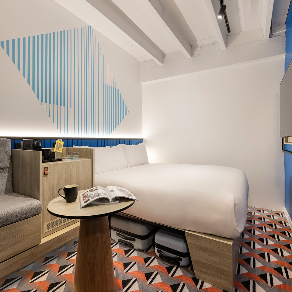 Modern hotel room with minimalist design. Features a geometric wall pattern, cozy bed, open magazine on round table, and vibrant patterned carpet.