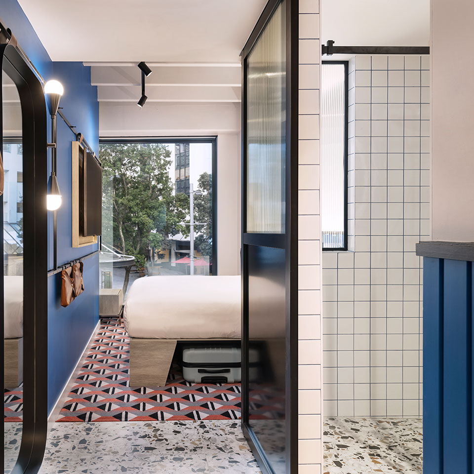 Stylish hotel room with blue walls, geometric red and black patterned floor, and large window overlooking trees. Modern, fresh, and urban ambiance.