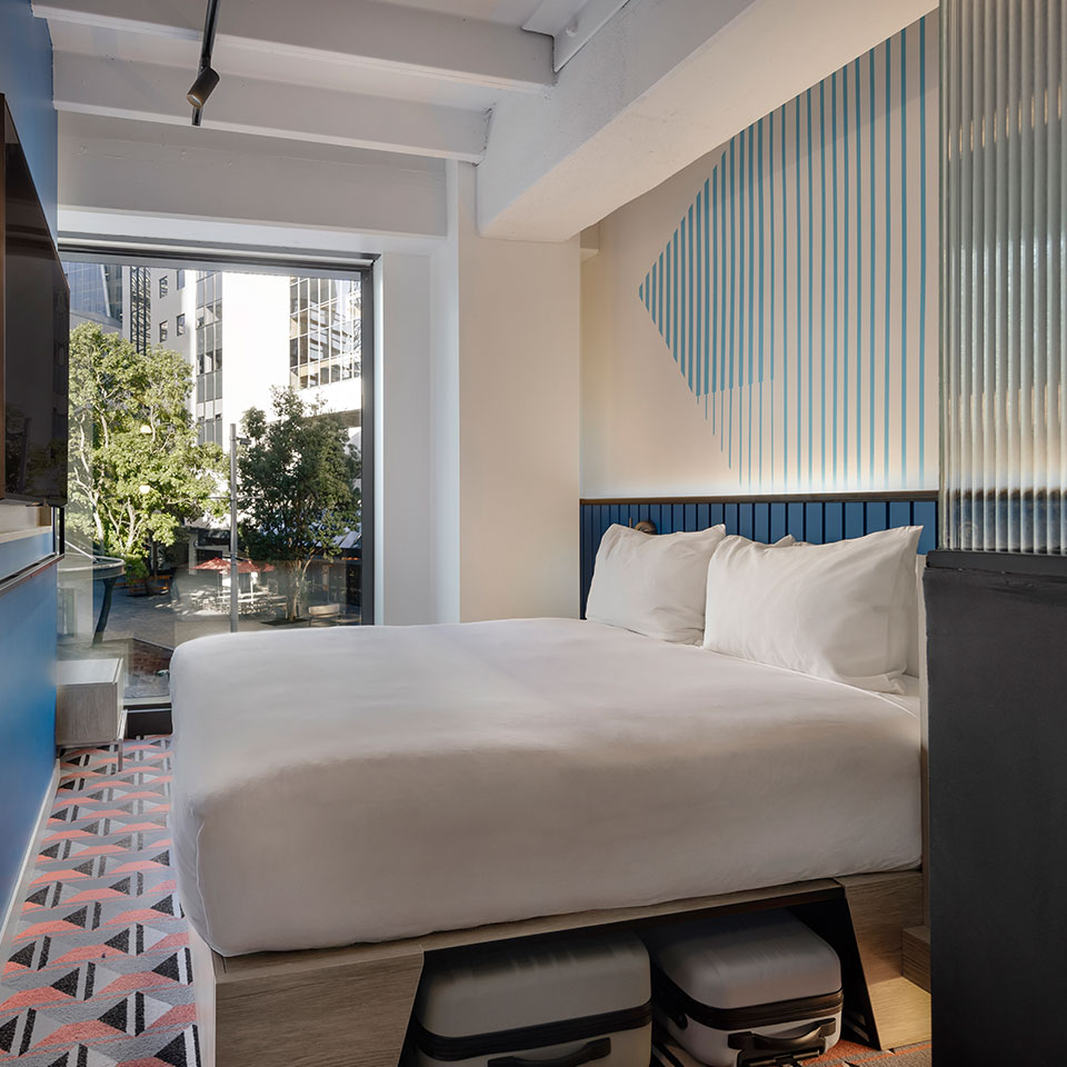 Modern hotel room with bold colors. Blue walls, geometric floor tiles, glass partition, and white bedding create a vibrant and cozy atmosphere. A large window shows trees outside.