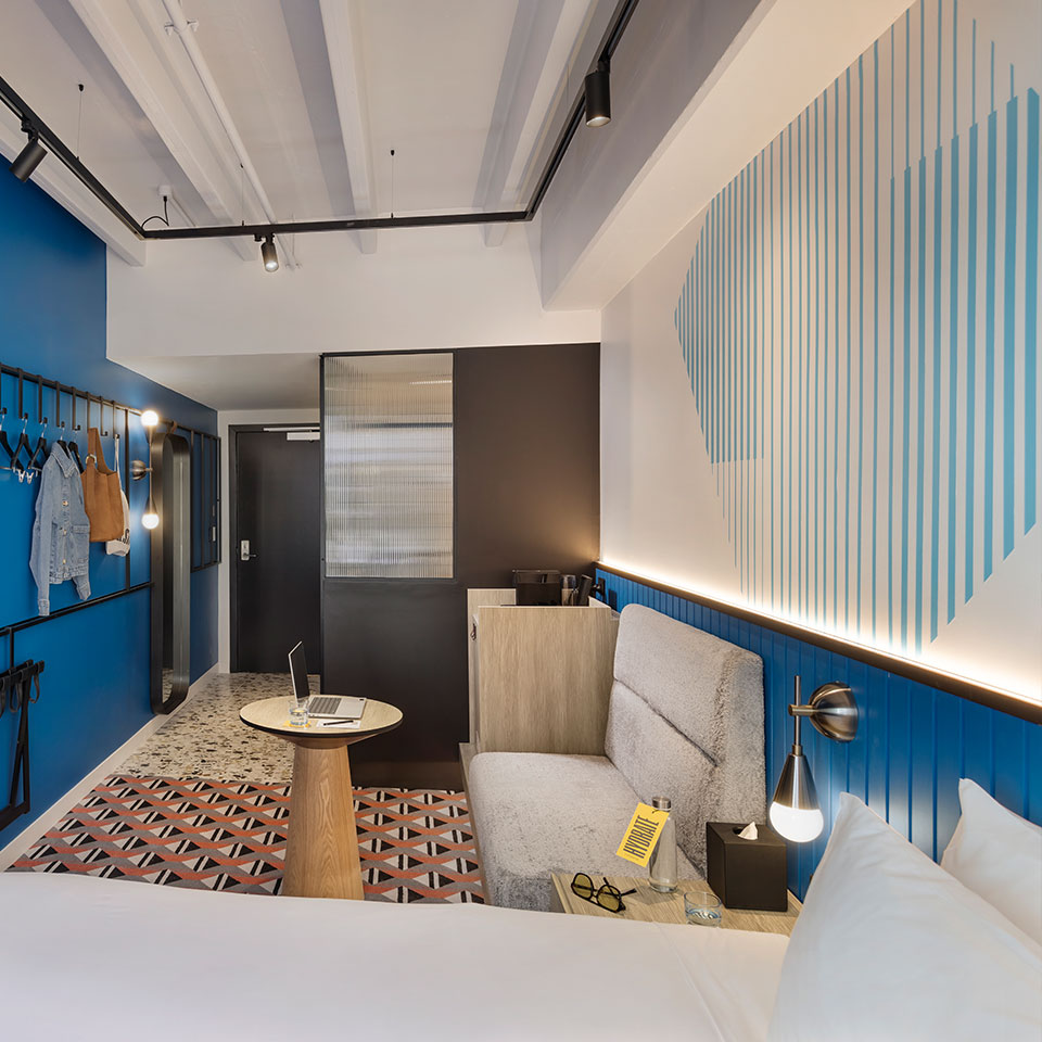Modern hotel room with blue accents and geometric patterns. Features a cozy bed, small sofa, round table with a laptop, and wall art. Bright and tidy ambiance.