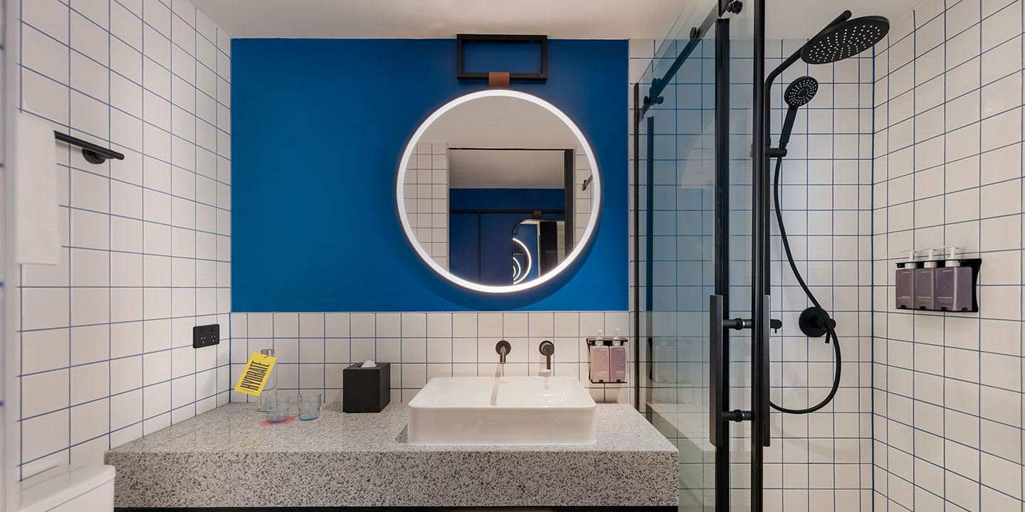 Modern bathroom with a circular backlit mirror on a blue wall above a rectangular sink. Black shower fixtures and white tiles create a sleek, clean look.