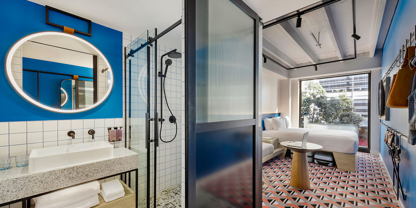 Modern hotel room with vibrant blue walls. Features include a circular mirror, open shower, double bed, and patterned red floor, exuding a stylish ambiance.