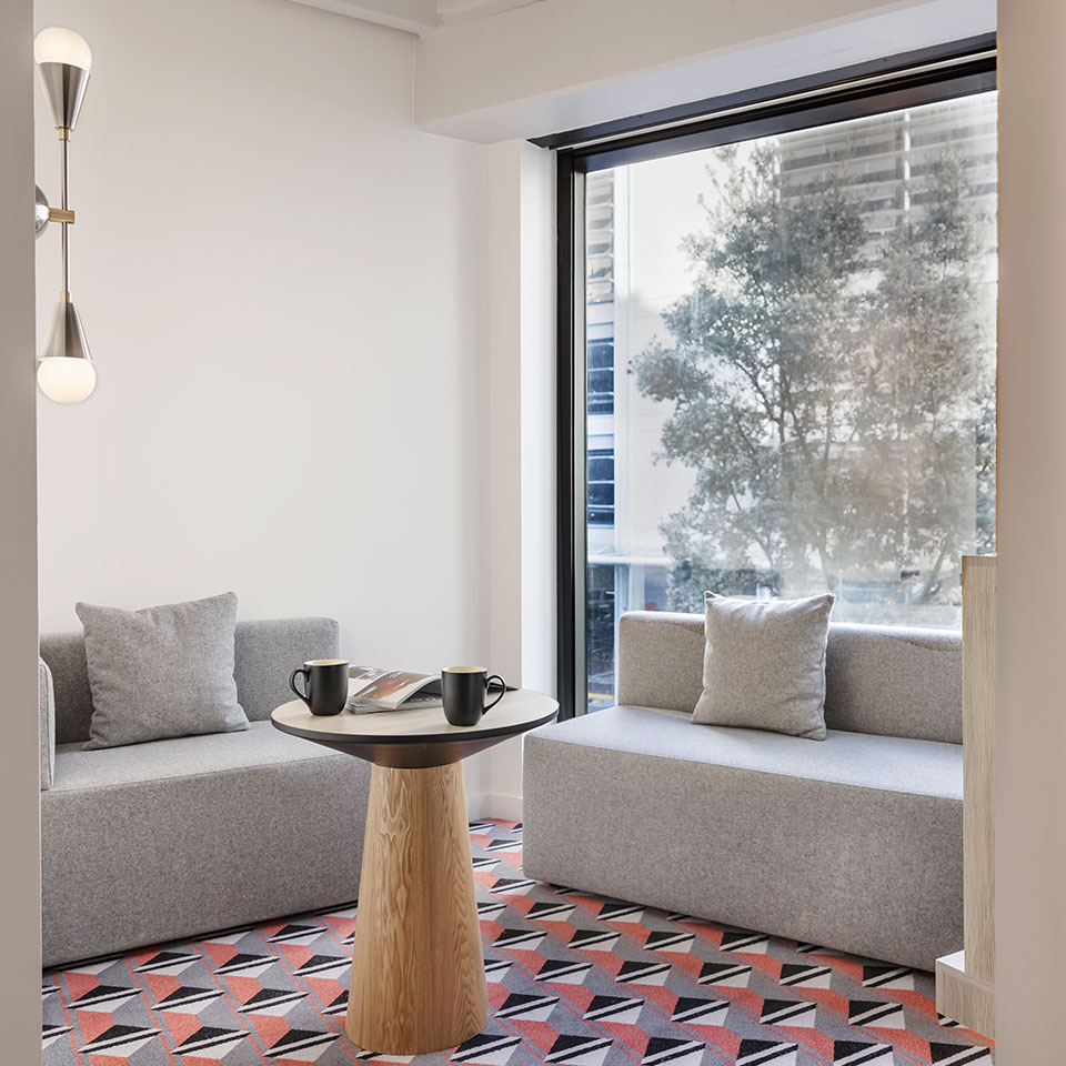 A bright, modern room with two gray sofas facing each other, a small wooden table in between with mugs and a magazine. Large window with trees outside.