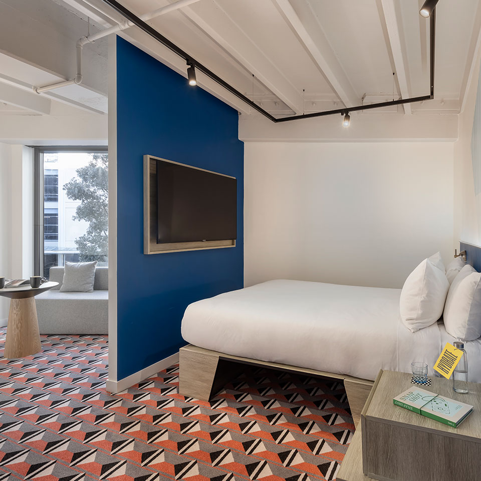 Modern hotel room with geometric patterned carpet, white bed against a blue accent wall, and flat-screen TV. A cozy sitting area is visible by the window.