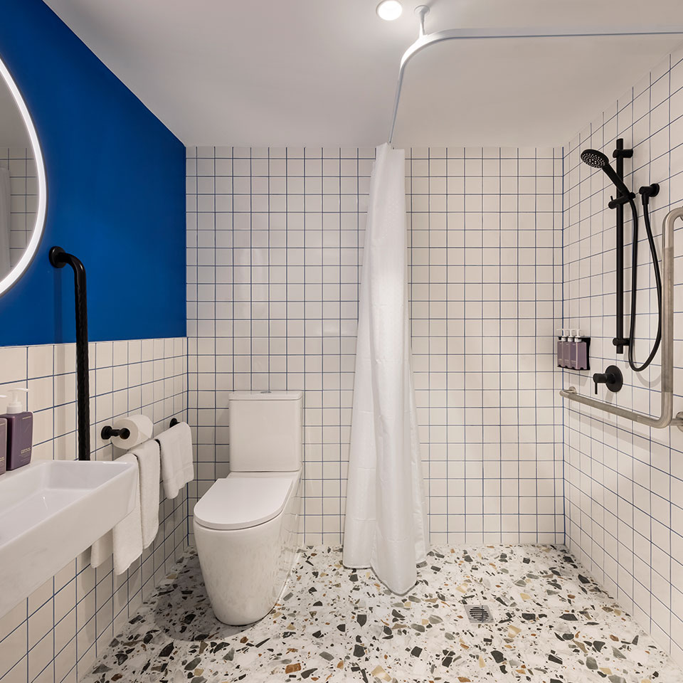 Modern bathroom with white square tiles, terrazzo flooring, and a navy blue accent wall. Features a round mirror, sleek fittings, and a white shower curtain.
