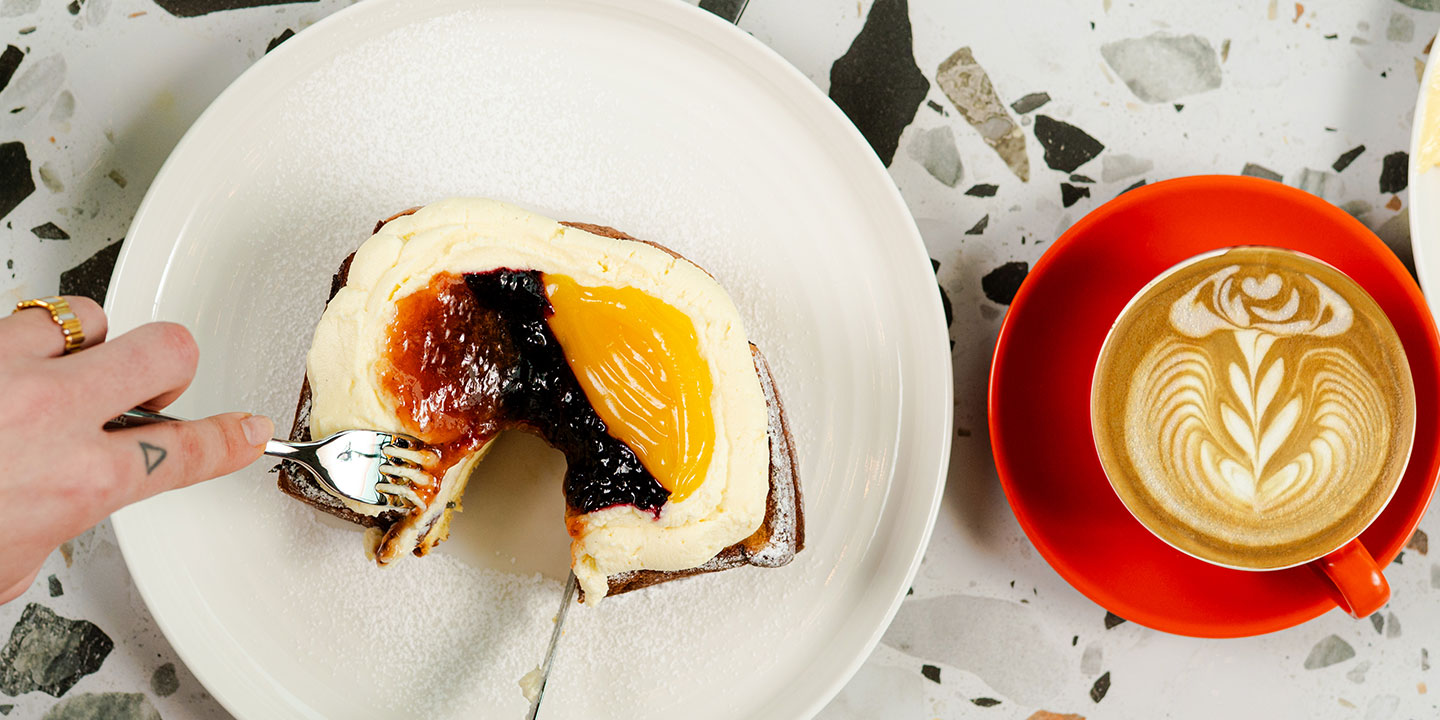 A hand with rings cuts into a pastry topped with red, yellow, and purple fruit jams on a white plate. Beside it, a latte with a leaf pattern sits on an orange saucer, set on a terrazzo surface.
