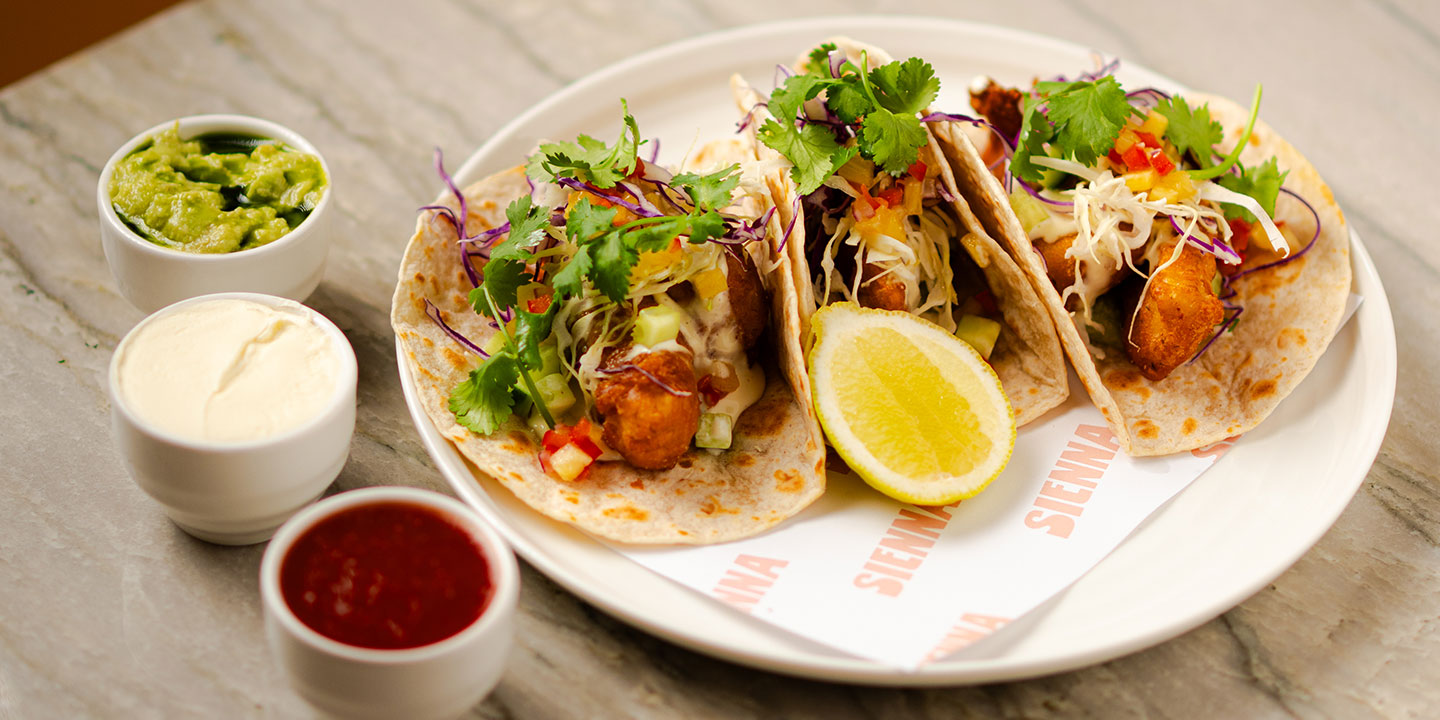 Three fish tacos on a plate with cilantro, red cabbage, and lemon. Sides of guacamole, sour cream, and salsa add vibrant color to a casual dining scene.