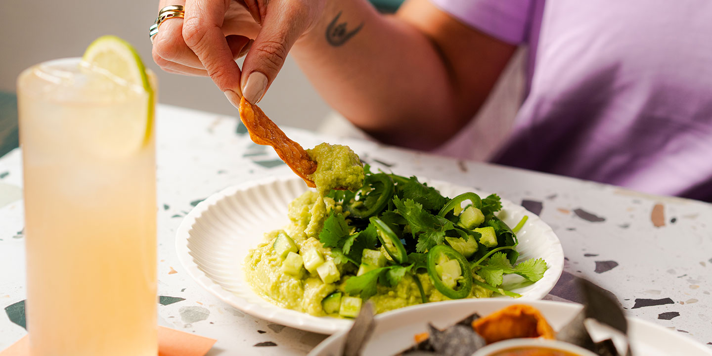 A person dips a chip into guacamole topped with cilantro and jalapeño on a terrazzo table. A refreshing drink with a lemon slice is nearby. Casual and inviting setting.