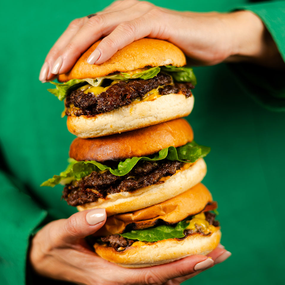 A person in a green shirt holds a stack of three juicy cheeseburgers with lettuce, emphasizing abundance and deliciousness.