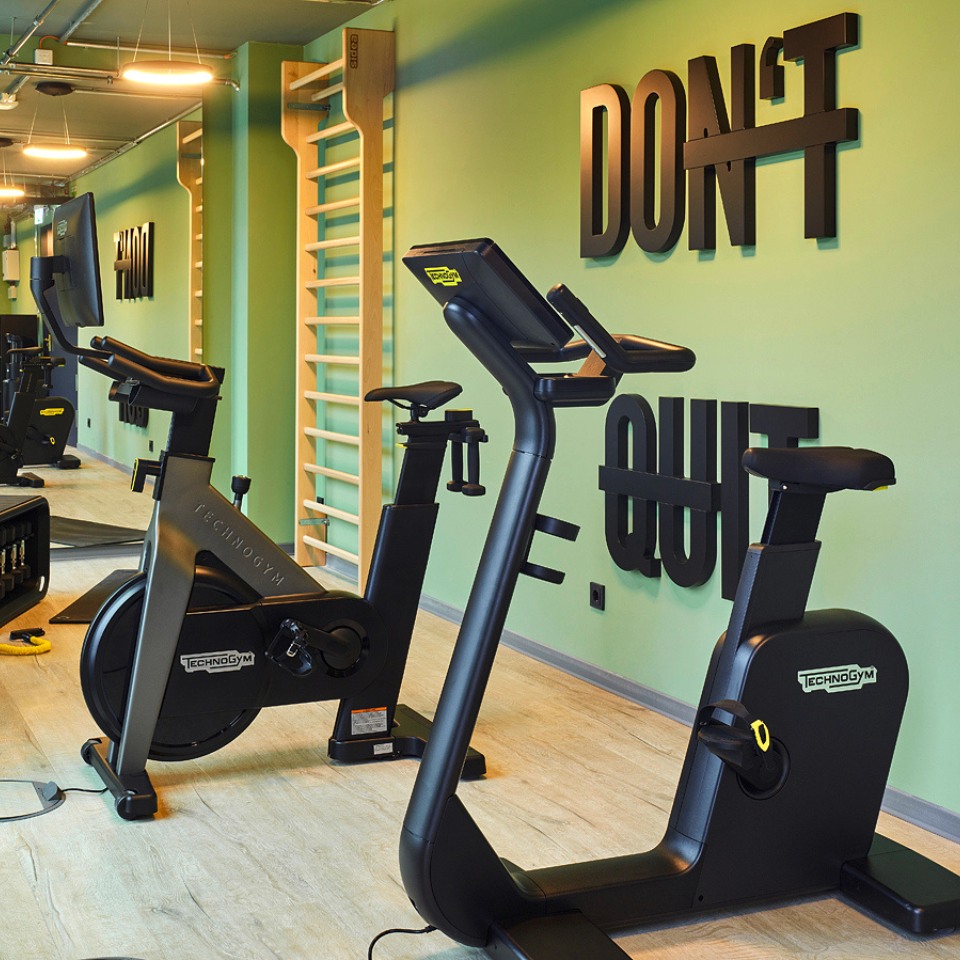 A modern gym interior featuring Technogym exercise bikes, a wooden climbing frame, and motivational wall art.