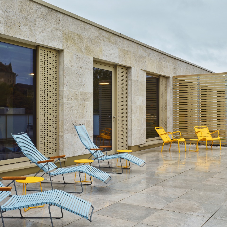 Terrace featuring striped blue lounge chairs and yellow metal chairs, surrounded by a stone wall and large windows.