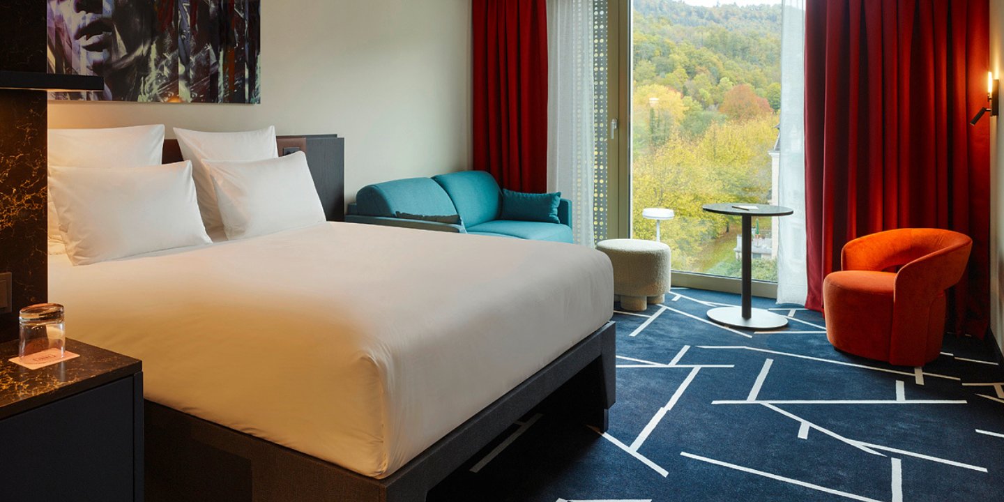 A hotel room featuring a large double bed, a stylish sofa, colorful decor, and a window view of lush greenery.