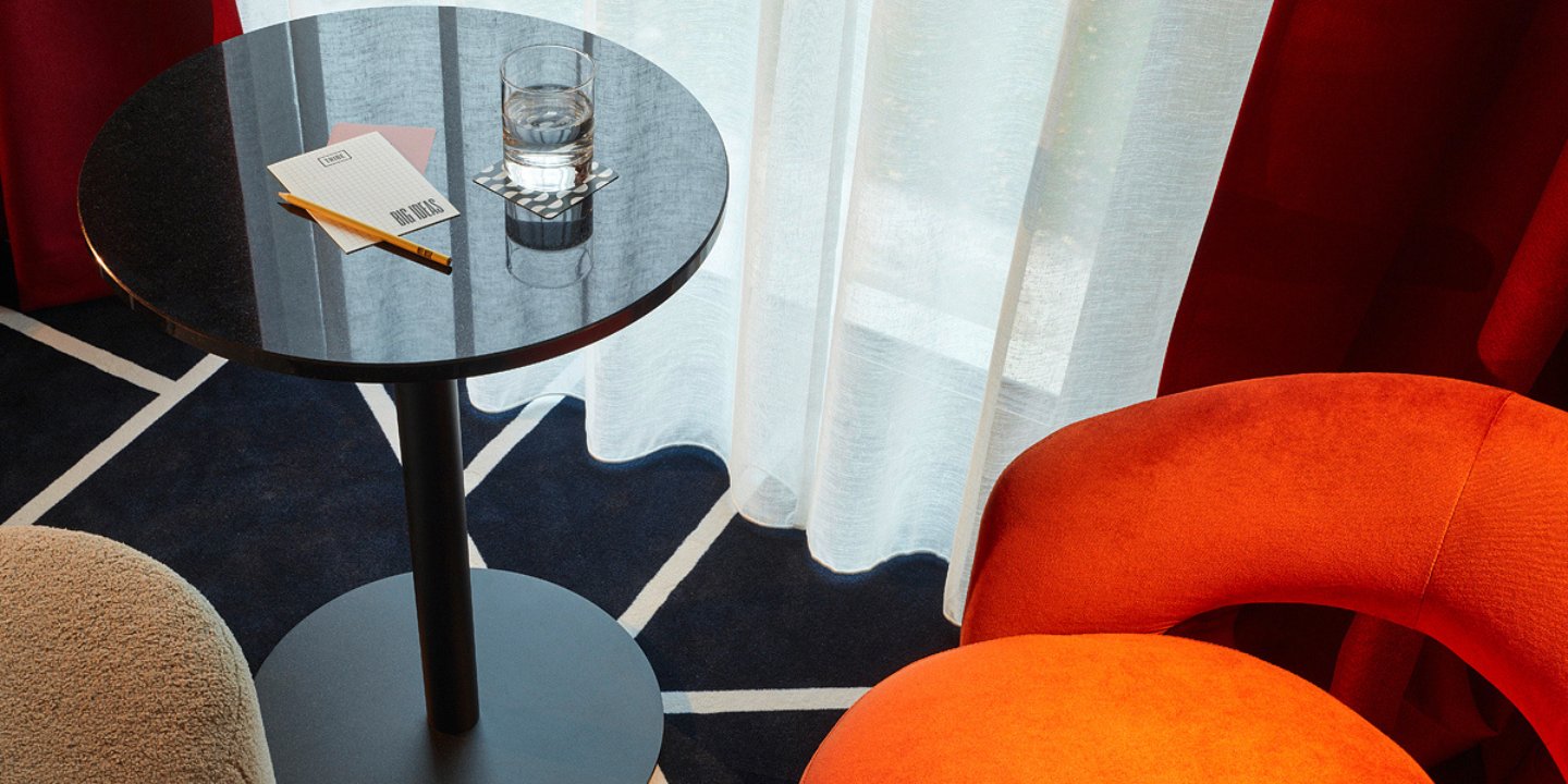 A modern table holds a glass of water and stationery, next to vibrant orange and plush chairs, set against soft curtains and a patterned rug.