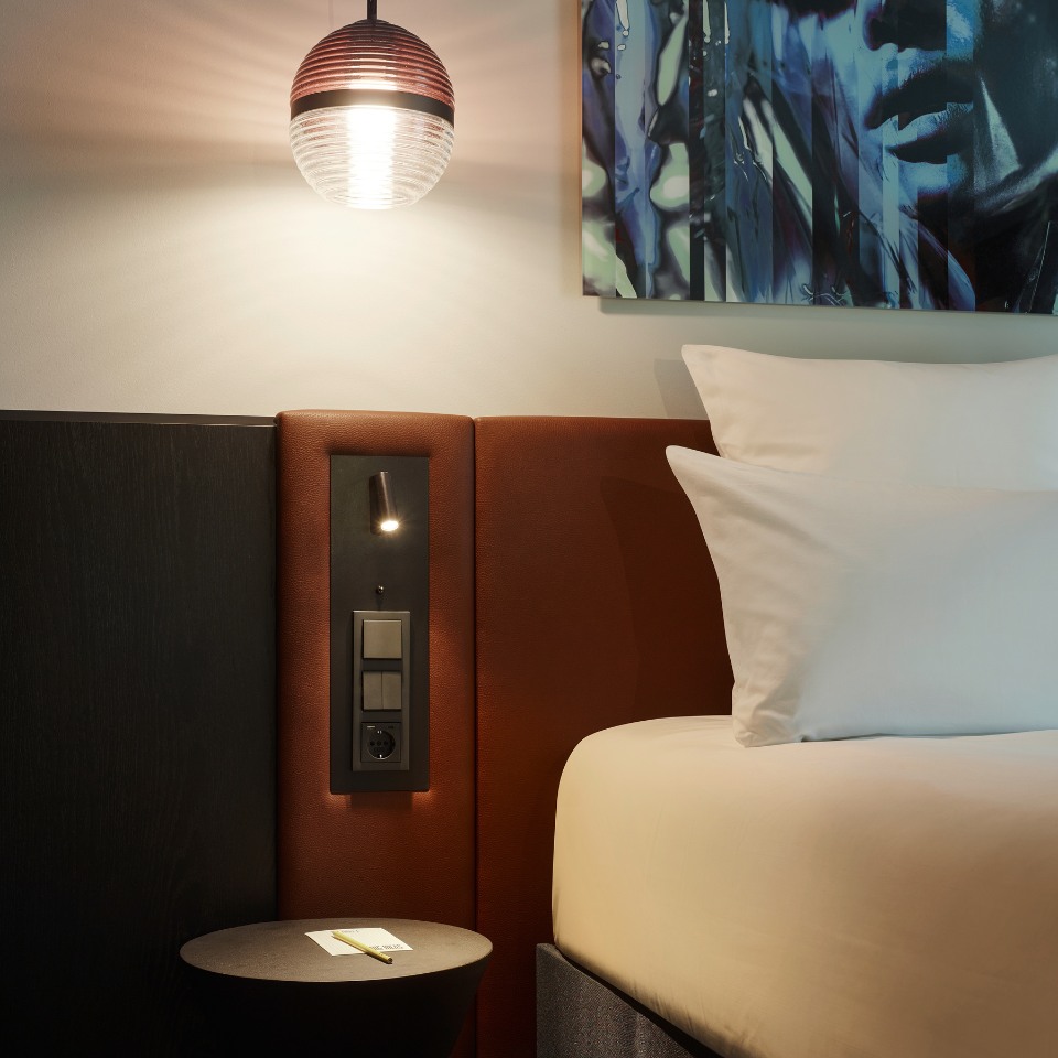 Cozy hotel room corner featuring a modern pendant light, plush bedding, and abstract wall art in warm tones.