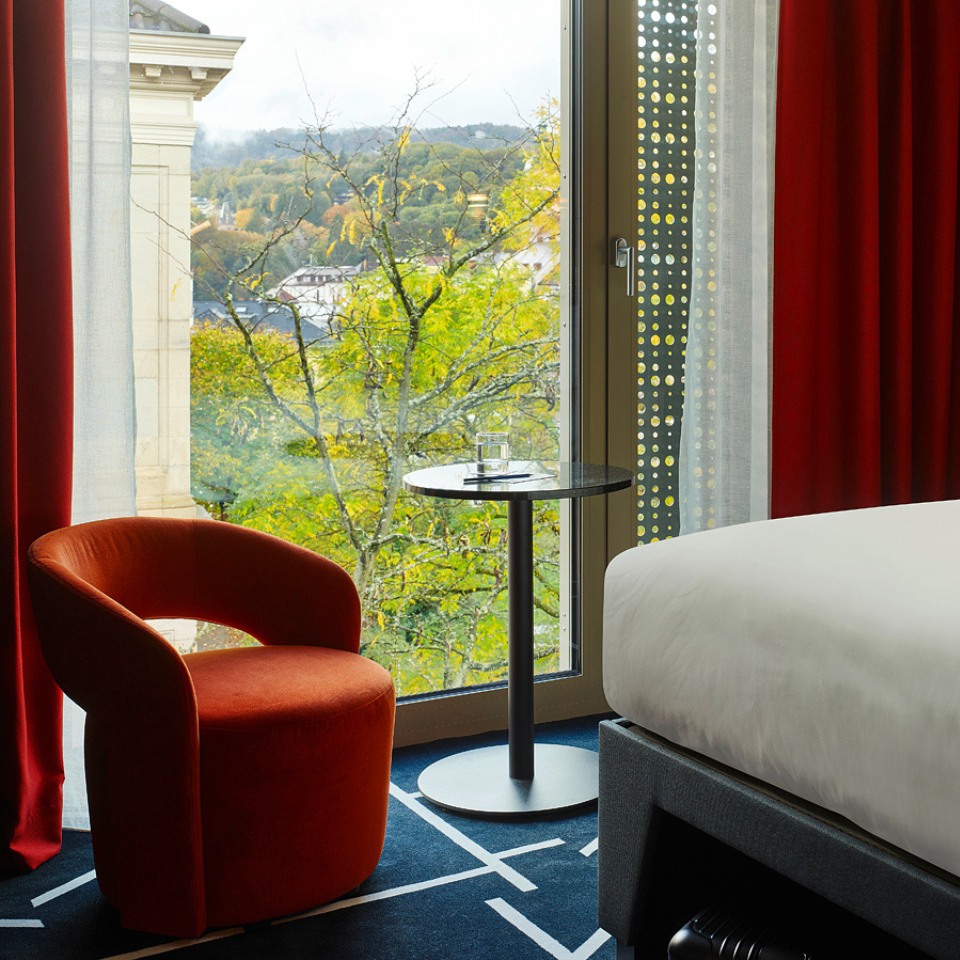 Cozy hotel room with a large window view of lush green trees, featuring a bed, orange chair, and minimalist decor.