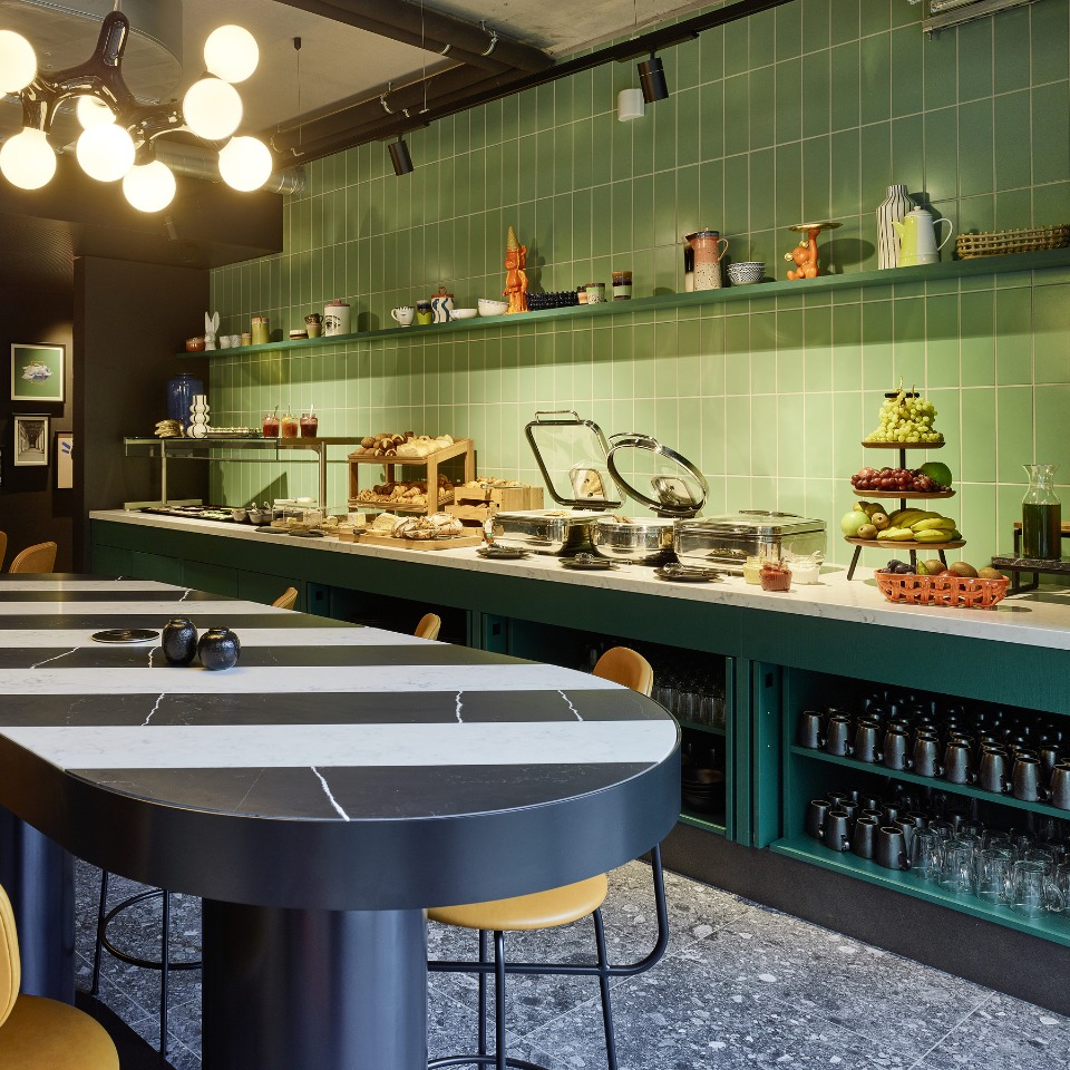 Breakfast setup with a green tiled wall, showcasing a buffet of pastries and fruit. There is an elegant table with yellow chairs.