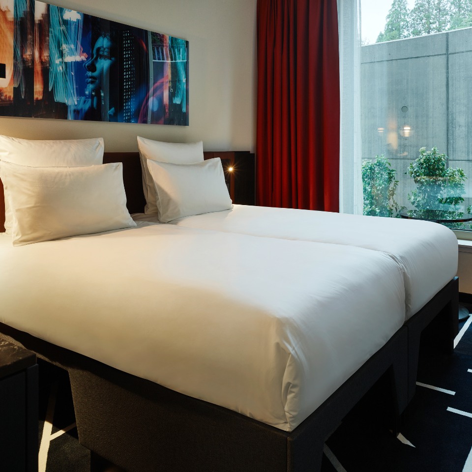 A modern hotel room featuring two neatly made beds with white linens, a colorful wall art piece, and red curtains with a garden view outside.