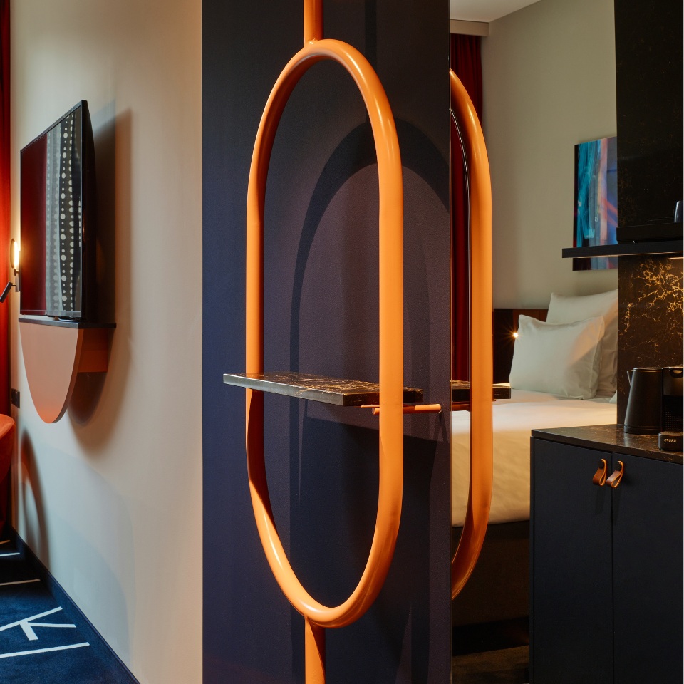 Hotel room featuring a unique orange archway with a shelf, minimalist decor, and a bed visible in the background.