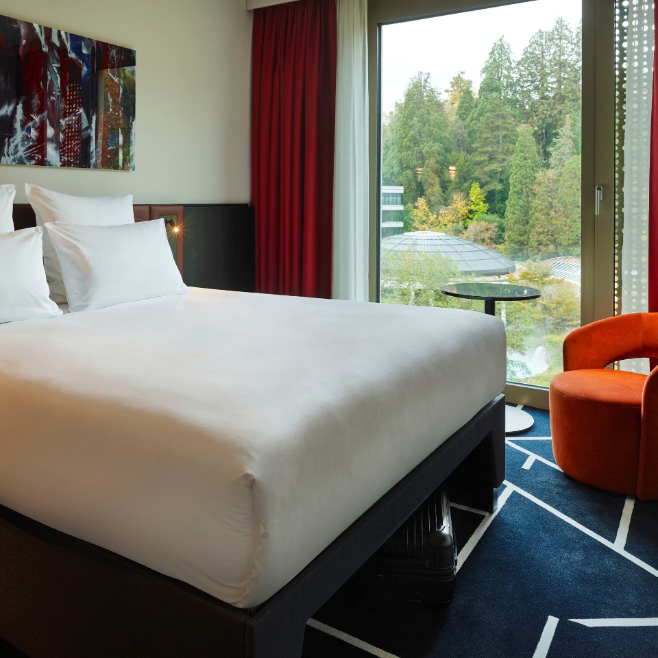 a double bed in a hotel room, the floor lenght windows show a forest, there is an orange armchiar in the corner