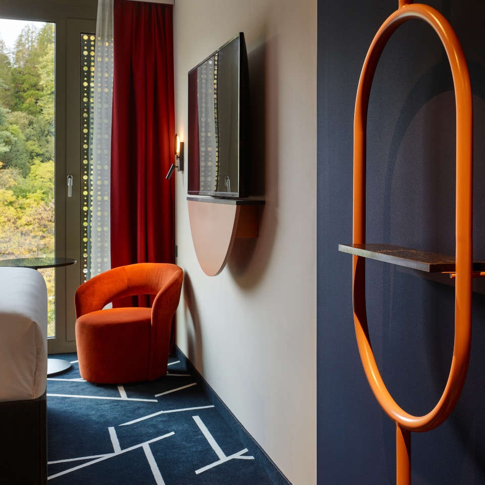 Hotel room featuring an orange chair, wall-mounted TV, and stylish decor, with red curtains framing a view of greenery outside.