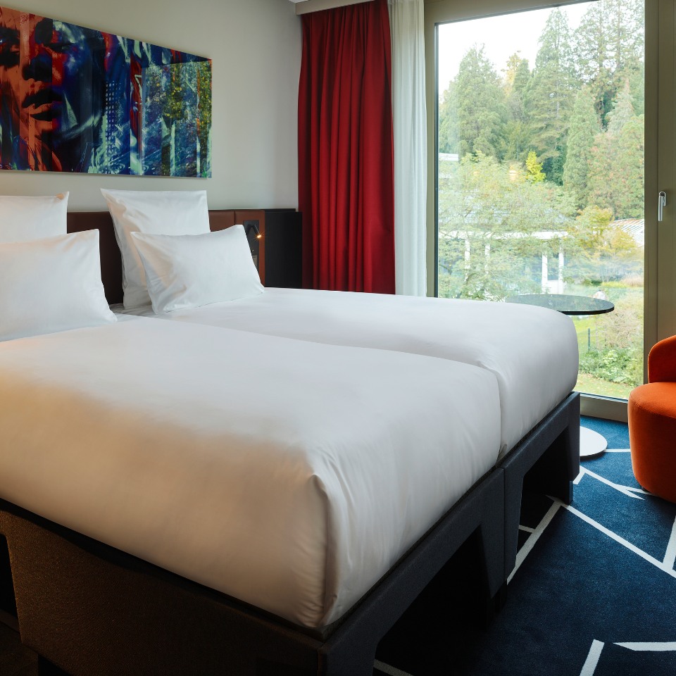 A modern hotel room featuring two beds, colorful artwork, and large windows overlooking lush greenery.