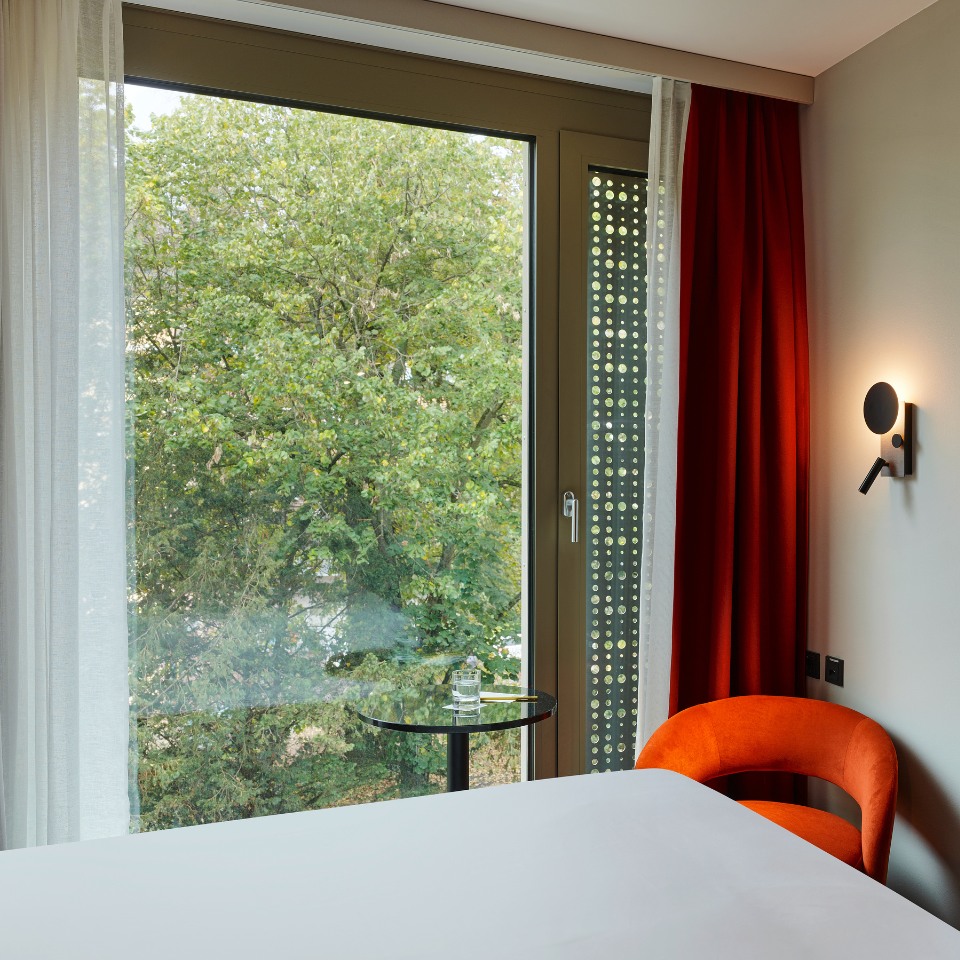 Cozy hotel room with a large window view of lush green trees, featuring a bed, orange chair, and minimalist decor.