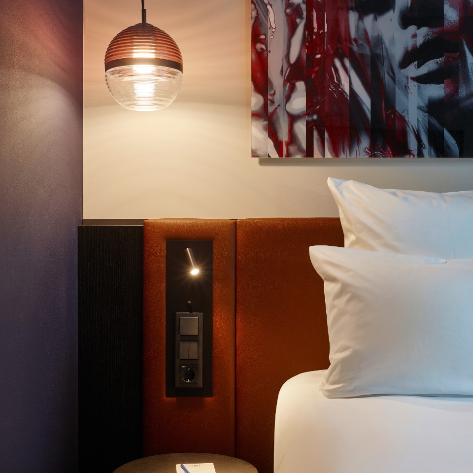 Cozy hotel room corner featuring a modern pendant light, plush bedding, and abstract wall art in warm tones.