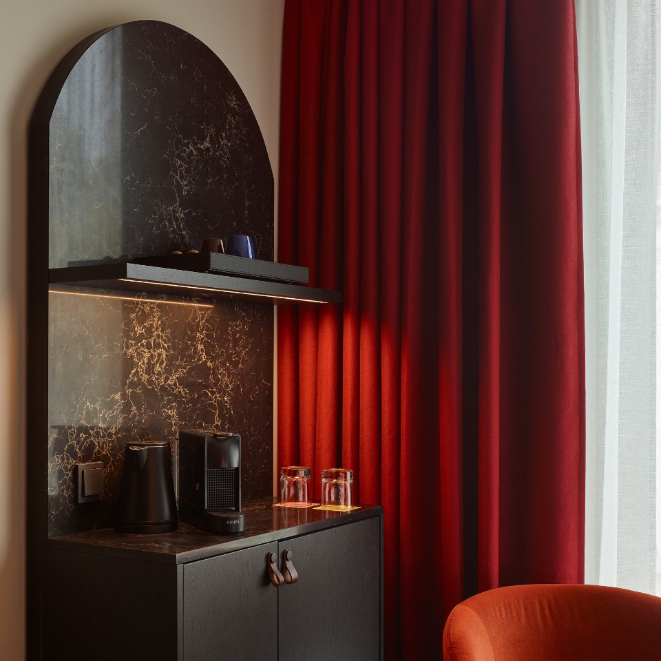 Hotel corner featuring dark marble shelf, coffee maker, glassware, and rich red curtains beside a cozy orange chair.
