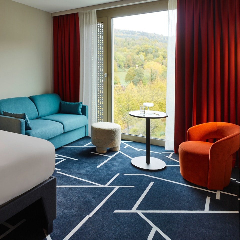 Hotel room with a teal sofa, orange chair, round table, and large window showcasing a scenic outdoor view.