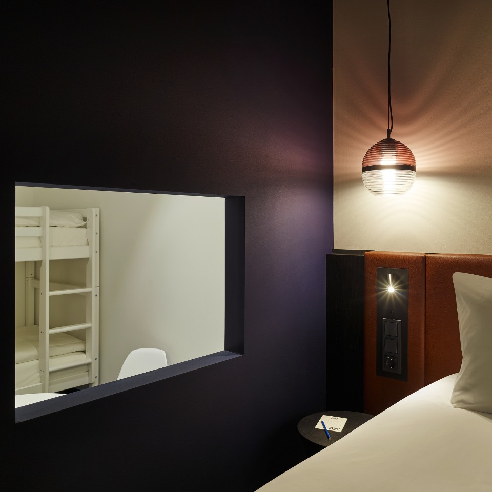 Hotel room featuring a bunk bed visible through a window, stylish pendant light, and a bedside table with a note pad.