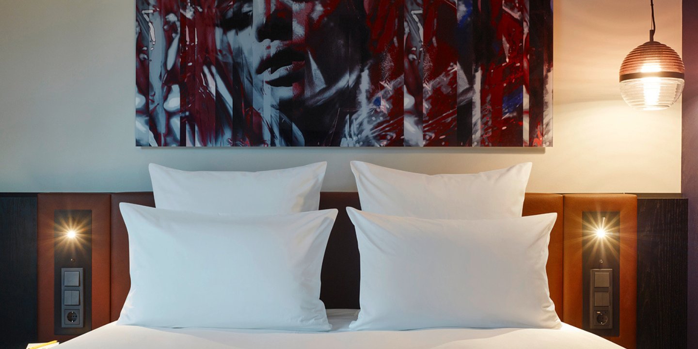 Hotel bedroom featuring a large artwork, white pillows on a brown bed, and stylish lighting fixtures on either side.