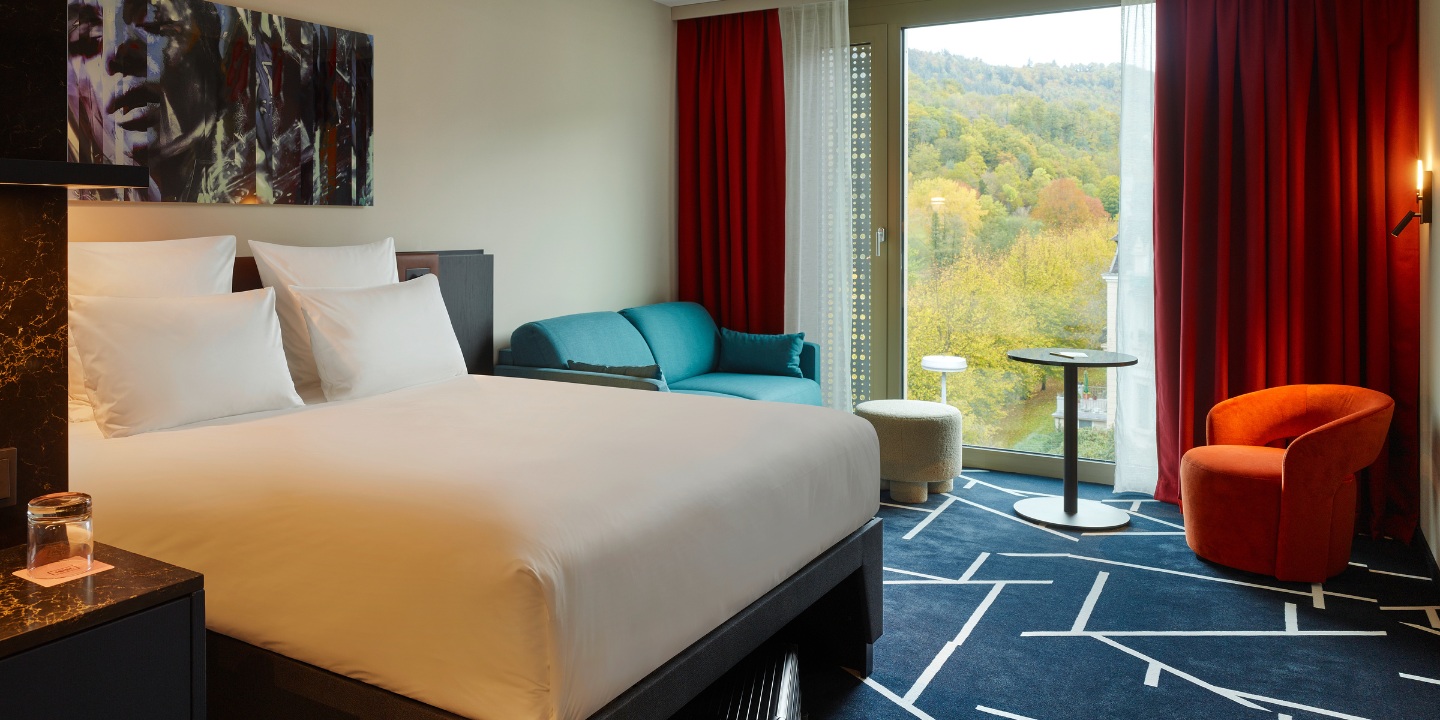Cozy hotel room featuring a large bed, colorful seating, and a balcony with scenic views of trees and hills outside.