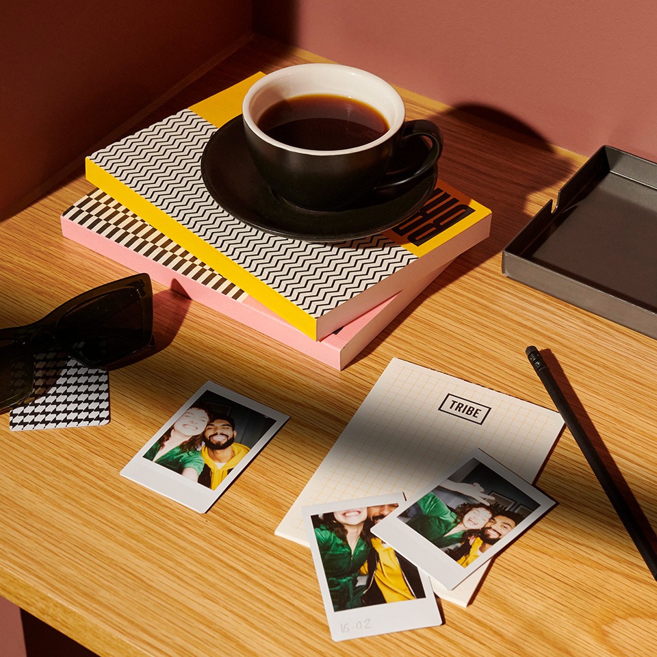 A black coffee cup sits atop colorful stacked notebooks on a wooden table, accompanied by sunglasses, photos, and a notebook with a pen.