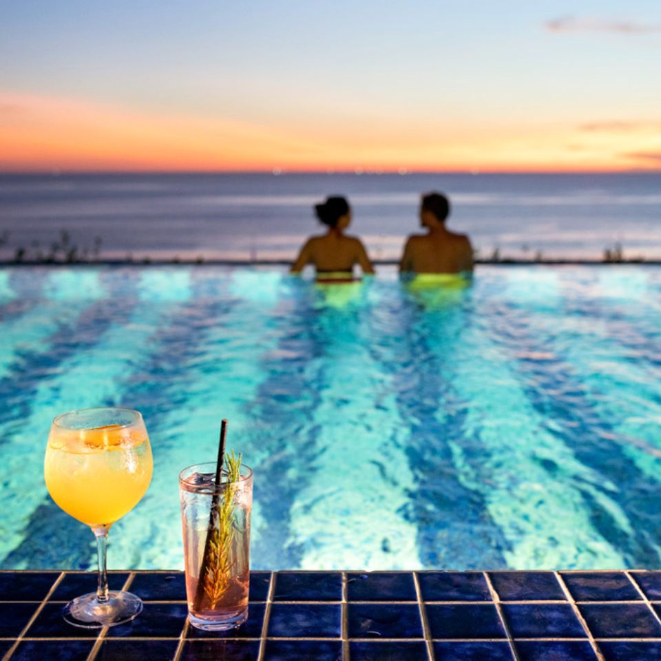 Two drinks on the side of a rooftop pool at sunset, there are two people swimming in the background