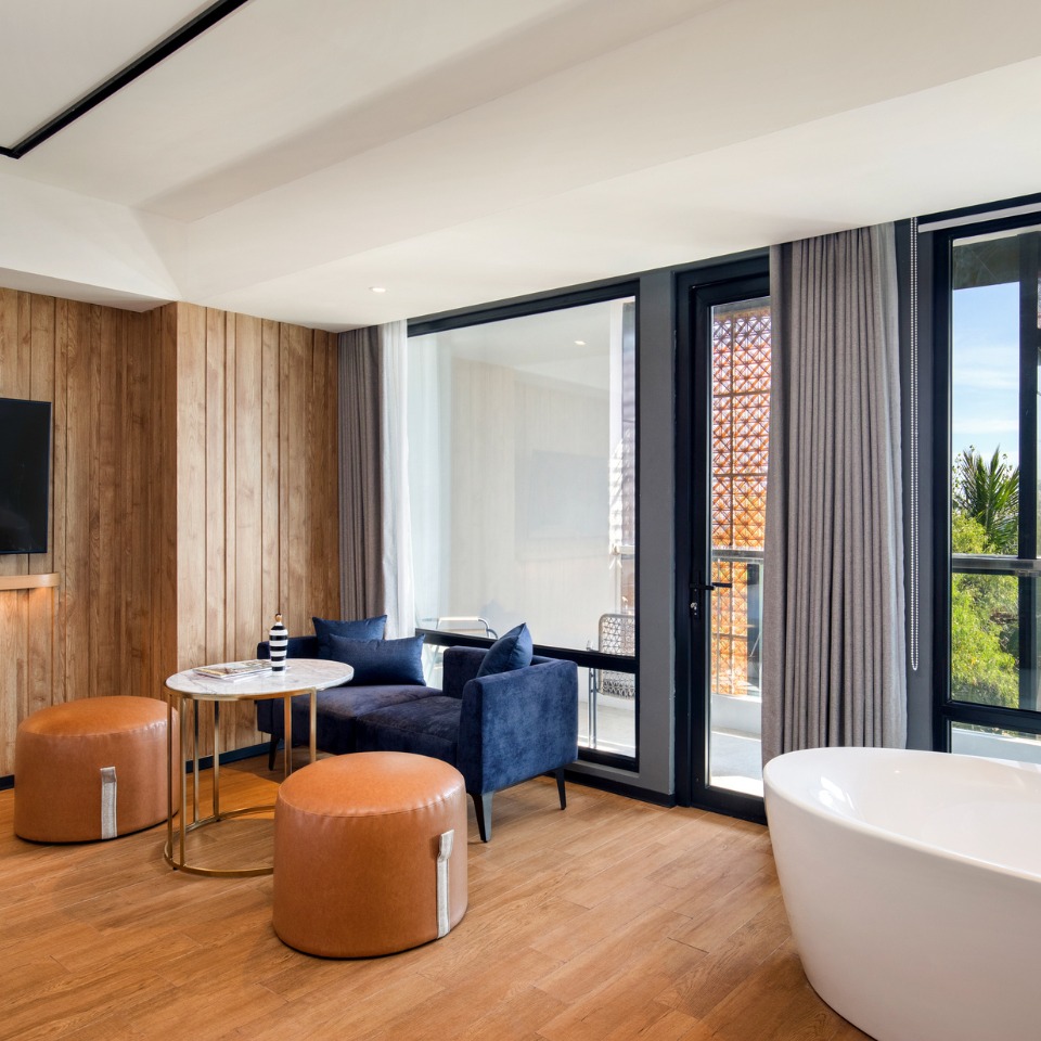 A hotel suite with a living area with a small blue couch and a white bathtub in front of a balcony