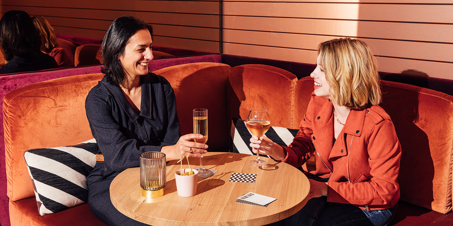 Two women enjoy drinks seated at a round wooden table adorned with snacks and napkins, surrounded by warm tones.