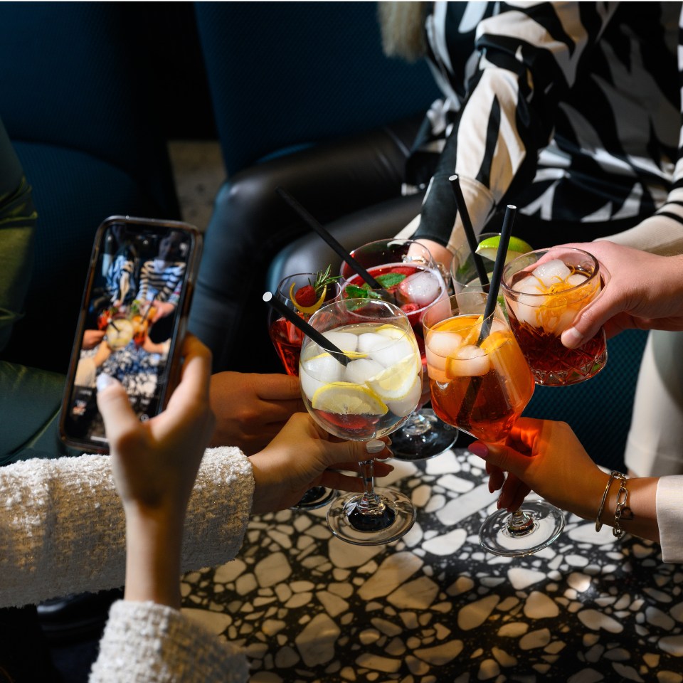 A group of friends cheers with colorful cocktails, sharing smiles and laughter, while one captures the moment on a smartphone.