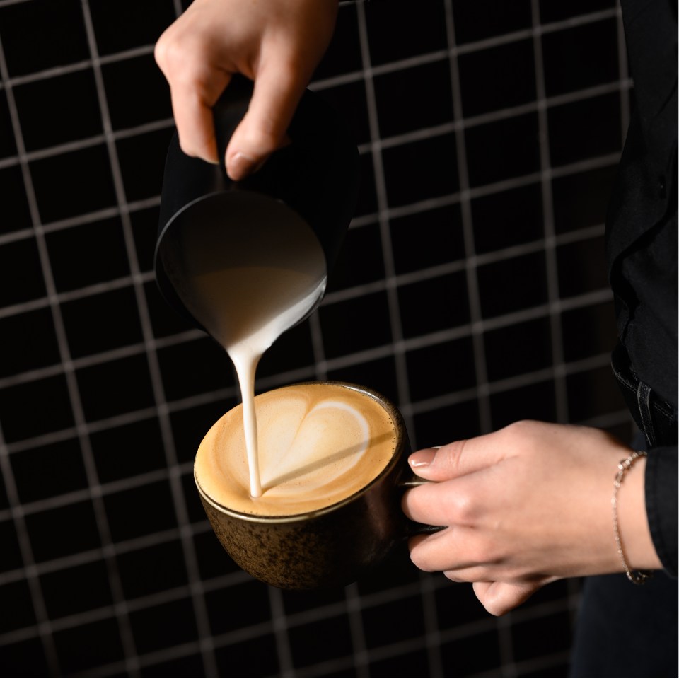 A barista pours creamy milk into a cup of coffee, creating a heart pattern on the surface against a black grid background.