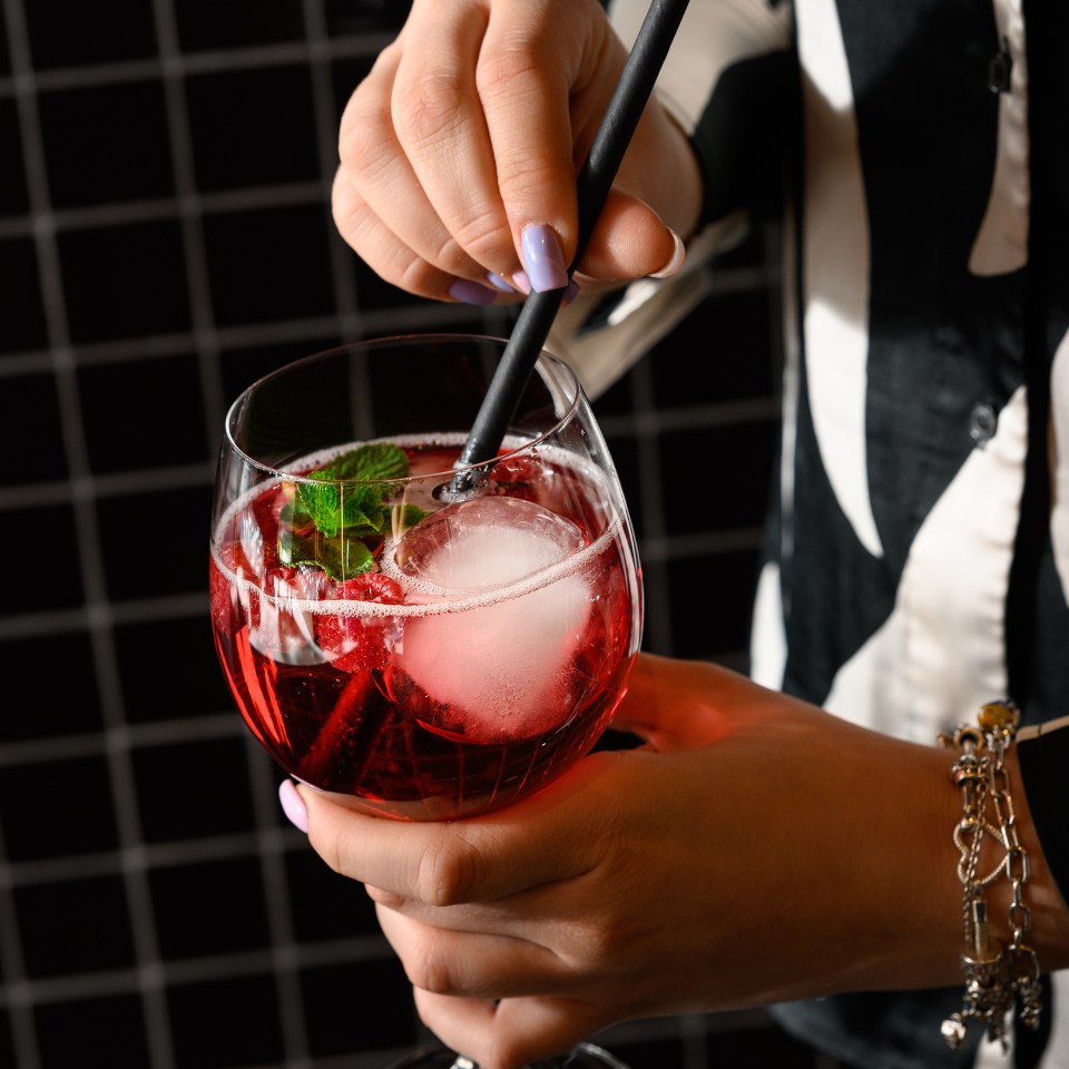 A hand stirring a vibrant red cocktail with mint and ice, using a black straw, against a patterned black and white background.