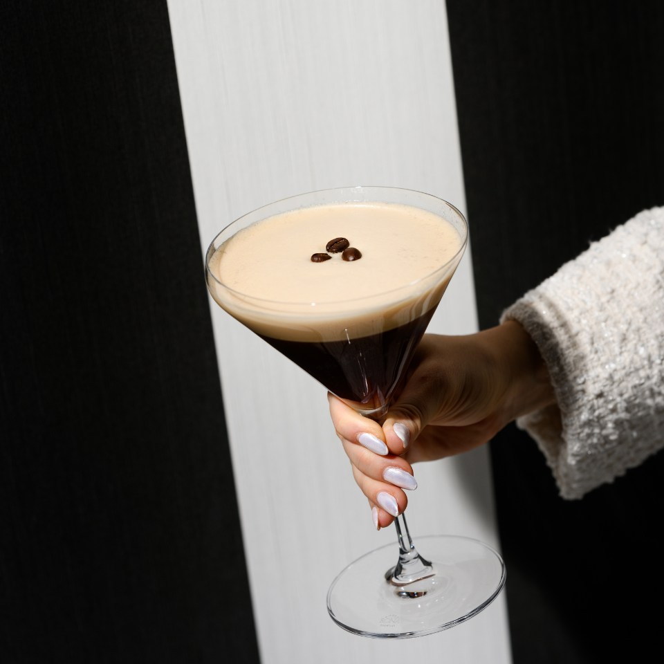 A hand holding a martini glass filled with an espresso martini, topped with three coffee beans, against a striped background.