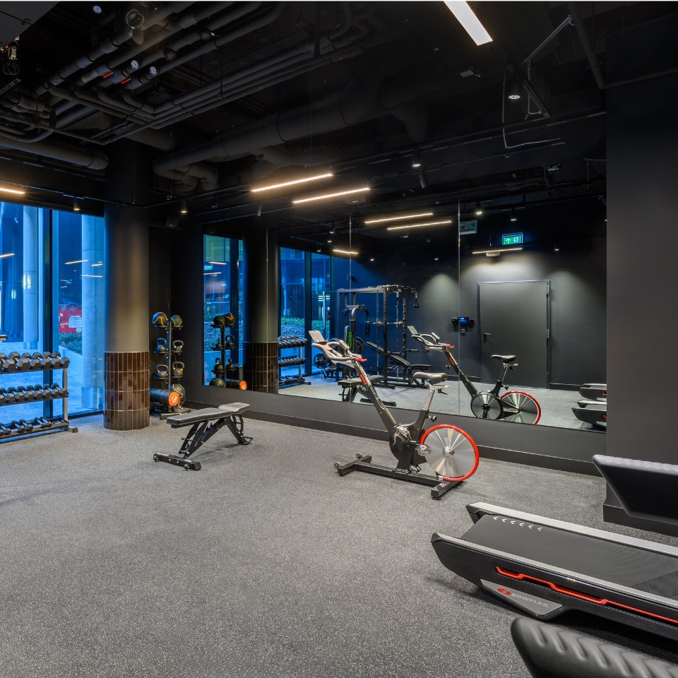 Gym interior featuring exercise bikes, weights, and benches, with large windows and mirrors reflecting the space.