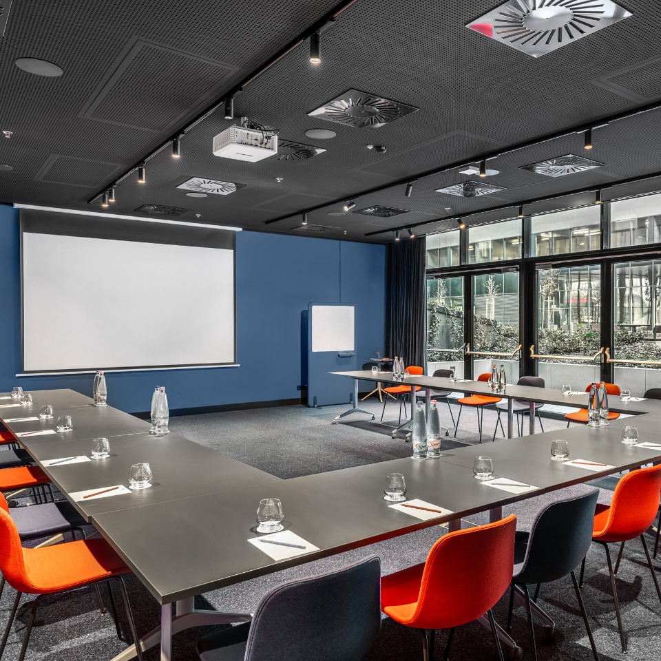 Modern conference room with a U-shaped table, orange chairs, projector screen, and large windows showcasing outdoor greenery.
