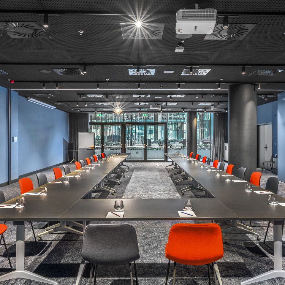 Modern conference room with long tables arranged in a U-shape, featuring colorful chairs and large windows for natural light.