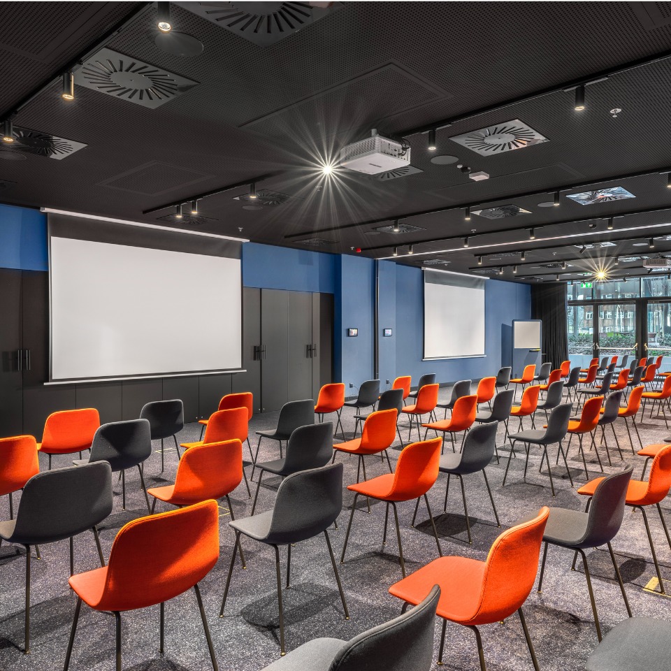 A large conference room with rows of black and orange chairs, two projection screens, and large windows for natural light.